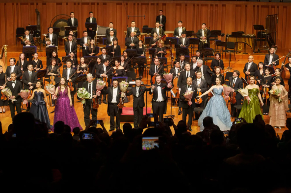 Dozens of orchestra musicians from the Philadelphia Orchestra and China National Symphony Orchestra stand on a stage in formal wear in front of an audience that is cheering and taking pictures of them.