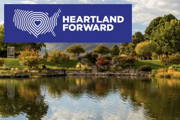 A serene park scene with a calm lake reflecting surrounding trees and hills. Above the landscape is a blue graphic with a white outline of the United States and the text Heartland Forward.