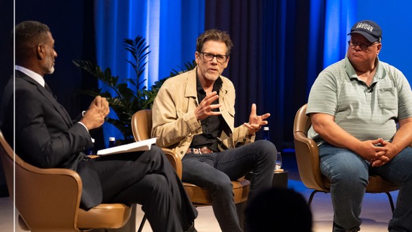 Three men are sitting in coversation in against a blue curtain background. The group includes Charles Blow, from left to right, in a dark suit and tie. The middle man, Kevin Bacon, wears glasses and a beige jacket over a dark shirt. The man on the right, Gabe Brown, wears a blue cap and a striped polo shirt.