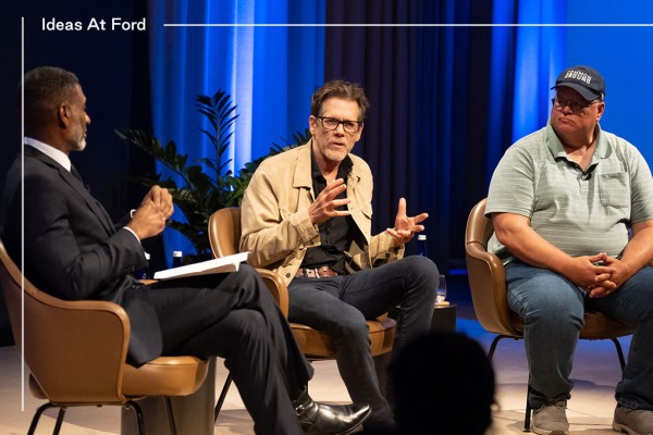 Three men are sitting in coversation in against a blue curtain background. The group includes Charles Blow, from left to right, in a dark suit and tie. The middle man, Kevin Bacon, wears glasses and a beige jacket over a dark shirt. The man on the right, Gabe Brown, wears a blue cap and a striped polo shirt.