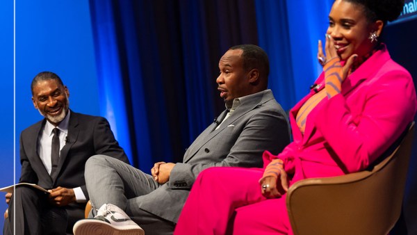 Three people are sitting in conversation against a blue curtain background. From left to right, they are Charles Blow in a dark suit and tie, Roy Wood Jr. in a grey suit and sneakers, and Joyelle Nicole Johnson in a bright pink suit.