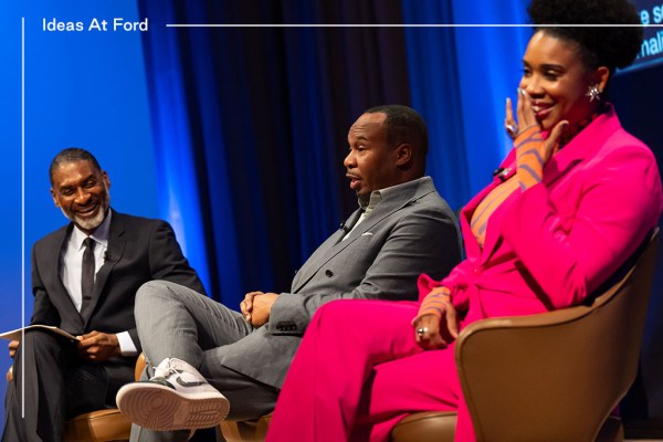 Three people are sitting in conversation against a blue curtain background. From left to right, they are Charles Blow in a dark suit and tie, Roy Wood Jr. in a grey suit and sneakers, and Joyelle Nicole Johnson in a bright pink suit.